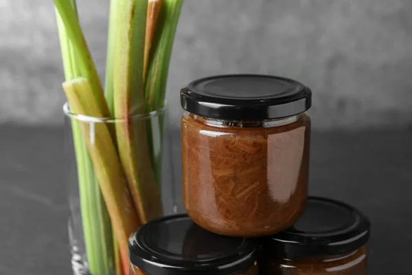 Jars of tasty rhubarb jam and stalks on grey table, closeup