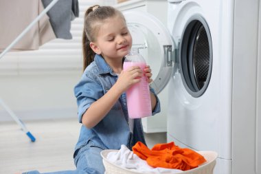 Küçük kız çamaşır makinesi ve sepetin yanında yumuşatıcı kokusu alıyor. Banyoda kirli giysiler, mesaj atmak için yer var.