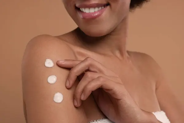 Young woman applying body cream onto arm on beige background, closeup