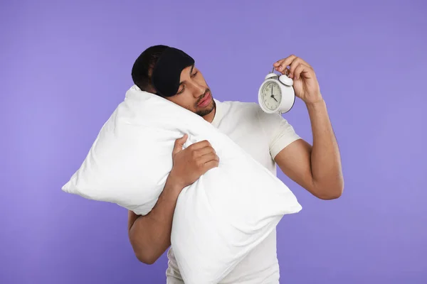 Tired man with pillow, sleep mask and alarm clock on violet background. Insomnia problem