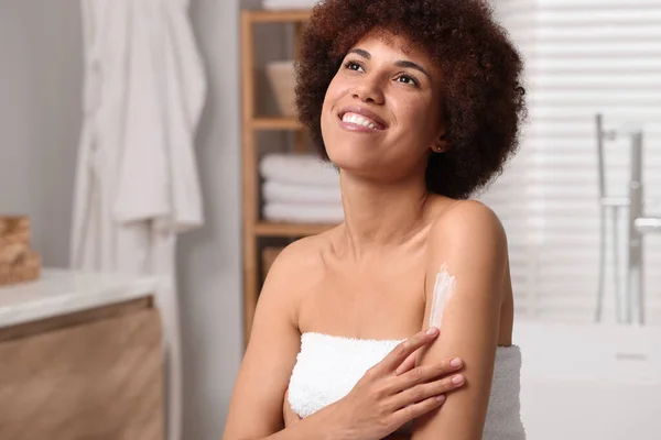 Beautiful young woman applying body cream onto arm in bathroom