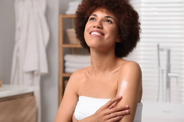 Beautiful young woman applying body cream onto arm in bathroom