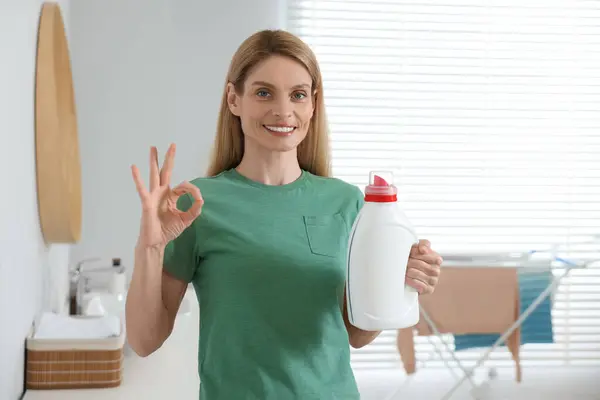 Woman holding fabric softener and showing OK gesture in bathroom