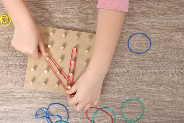 Motor skills development. Girl playing with geoboard and rubber bands at wooden table, top view