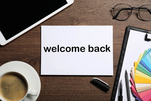 Paper with phrase Welcome Back on wooden table. Office desk with tablet and coffee, flat lay