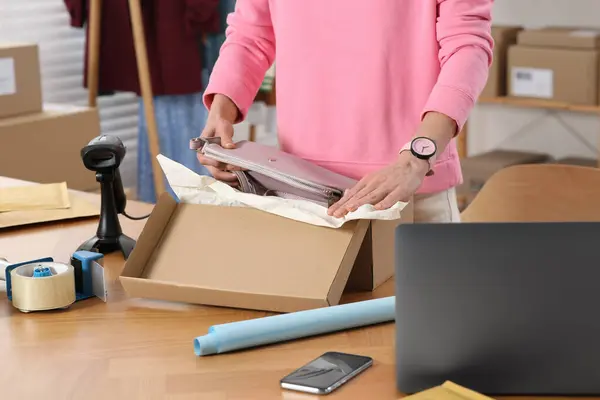 Seller packing bag into cardboard box at table in office, closeup. Online store