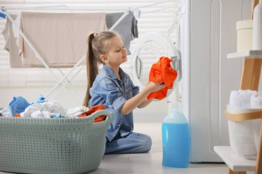 Küçük kız banyodaki çamaşır makinesine kirli çamaşır koyuyor.