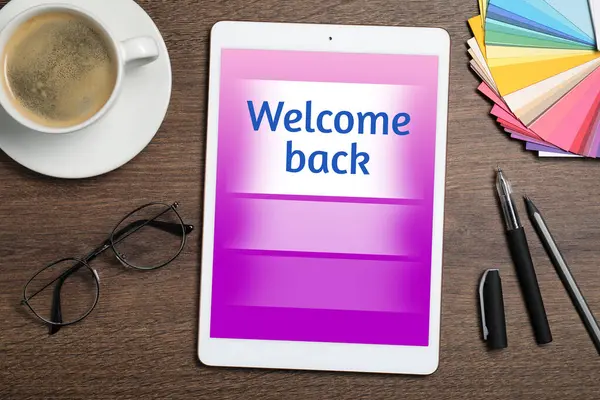 Tablet with phrase Welcome Back on wooden table. Office desk with cup of coffee, flat lay