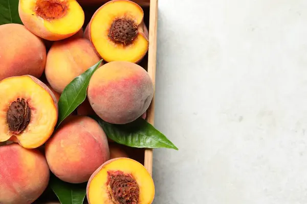 Delicious juicy peaches and leaves in crate on light grey textured table, top view. Space for text
