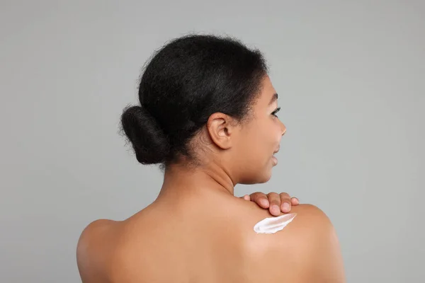 Young woman applying body cream onto back on grey background