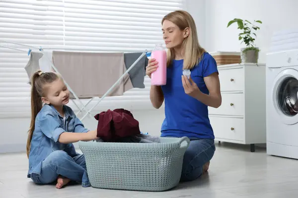 Mother smelling fabric softener while daughter taking out dirty clothes from basket in bathroom