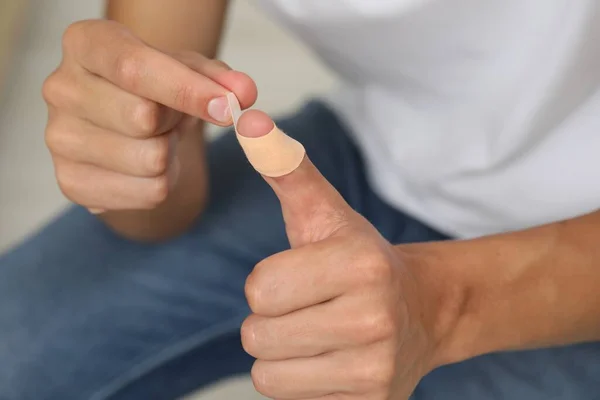 Man putting sticking plaster onto thumb indoors, closeup