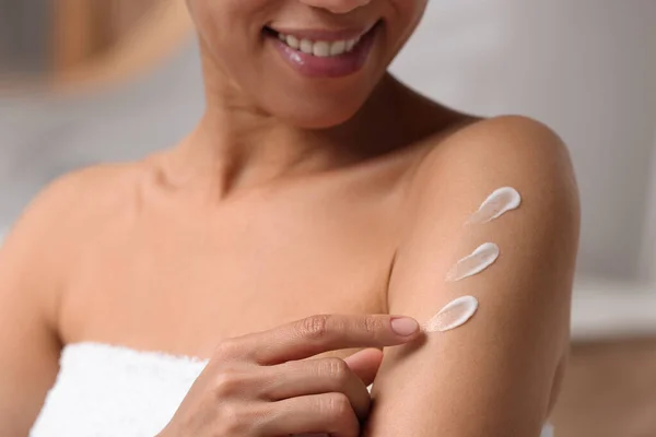 Beautiful young woman applying body cream onto arm on blurred background, closeup