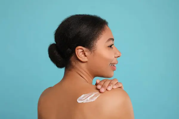 Young woman applying body cream onto back on light blue background