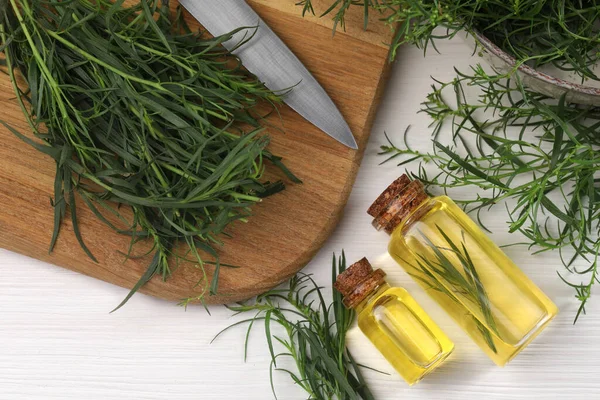 Bottles of essential oil and fresh tarragon sprigs on white wooden table, flat lay