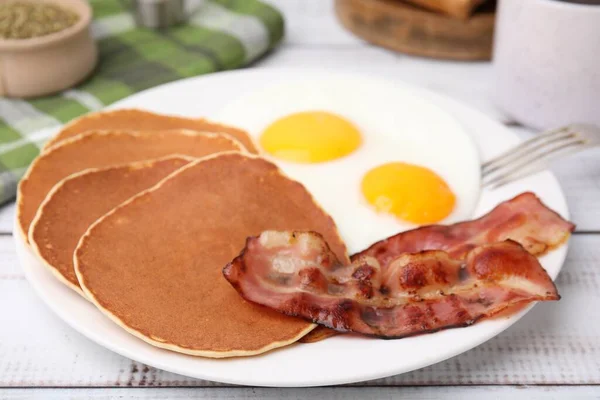 Tasty pancakes with fried eggs and bacon on white wooden table, closeup