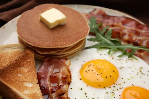 Tasty pancakes with fried eggs and bacon on plate, closeup