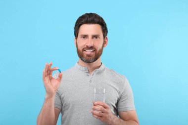 Açık mavi arka planda bir bardak su ve hap taşıyan yakışıklı bir adam.