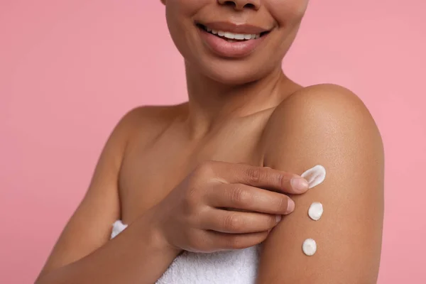 Young woman applying body cream onto arm on pink background, closeup