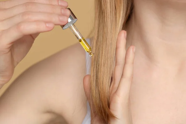 Woman applying essential oil onto hair on beige background, closeup