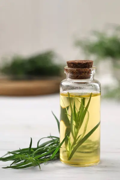 Bottle of essential oil and fresh tarragon leaves on white wooden table