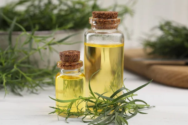 Bottles of essential oil and fresh tarragon leaves on white wooden table