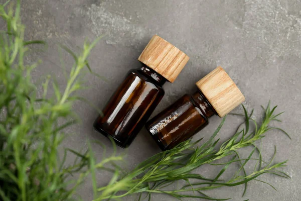 Bottles of essential oil and fresh tarragon leaves on grey table, flat lay