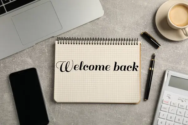 Notebook with phrase Welcome Back on grey table. Office desk with laptop and coffee, flat lay