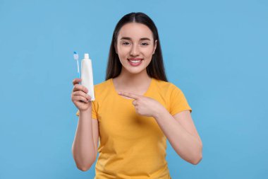 Açık mavi arka planda plastik diş fırçası ve tüp diş macunu tutan mutlu genç bir kadın.