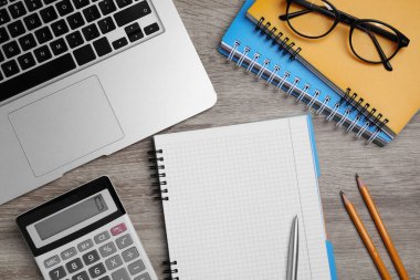 Defter, hesap makinesi, dizüstü bilgisayar, gözlük ve kırtasiye malzemesi ahşap masada, düz konum
