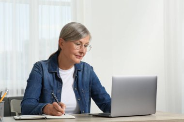 Güzel son sınıf öğrencisi not defterine notlar yazarken ahşap masada dizüstü bilgisayar kullanıyor.