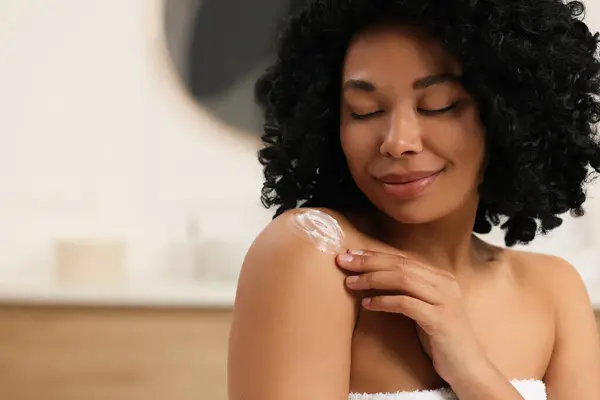 Young woman applying body cream onto shoulder indoors. Space for text