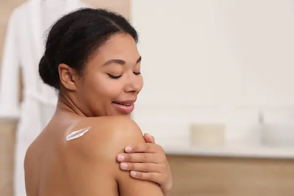 Young woman applying body cream onto back in bathroom. Space for text