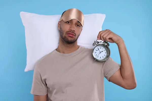 Tired man with pillow, sleep mask and alarm clock on light blue background. Insomnia problem