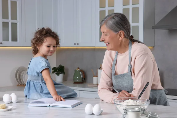 Cute little girl with her granny cooking by recipe book in kitchen