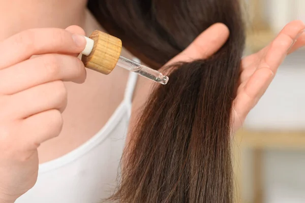 Woman applying essential oil onto hair on blurred background, closeup