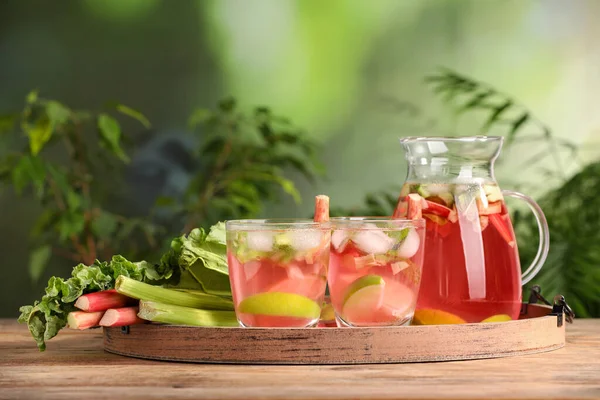 Glasses and jug of tasty rhubarb cocktail with citrus fruits on wooden table outdoors