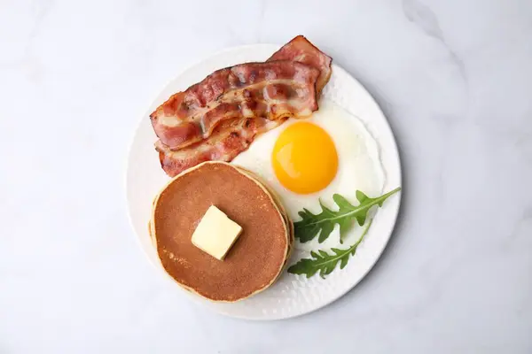 Tasty pancakes with fried egg and bacon on white table, top view