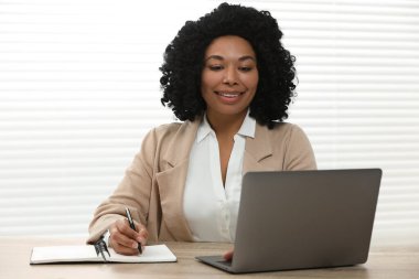 İçerideki ahşap masada dizüstü bilgisayar kullanırken notlar yazan mutlu genç bir kadın.