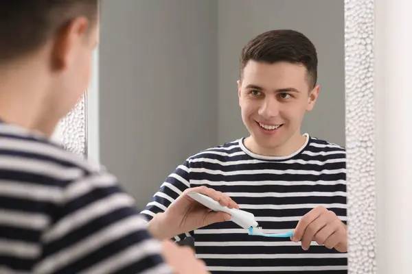 バスルームの鏡の近くの歯ブラシのチューブから歯磨き粉を絞る幸せな男 — ストック写真