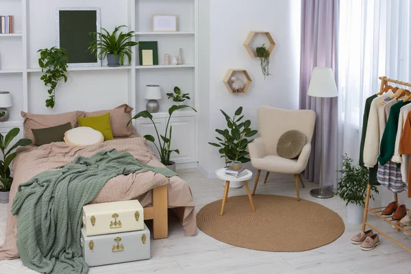 Stylish bedroom with comfortable bed, clothes rack and different houseplants. Interior design
