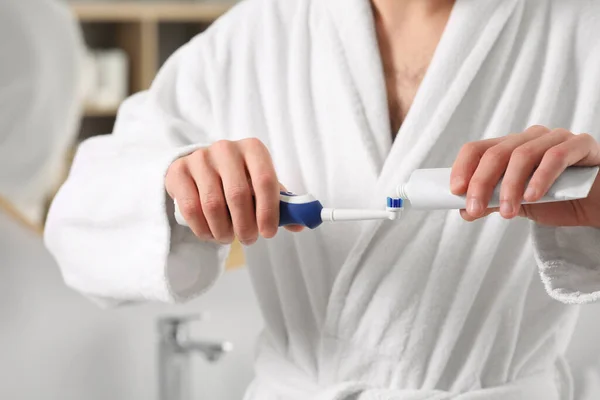 Man squeezing toothpaste from tube onto electric toothbrush indoors, closeup