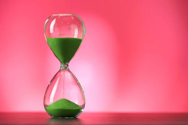 Hourglass with flowing green sand on table against pink background, space for text