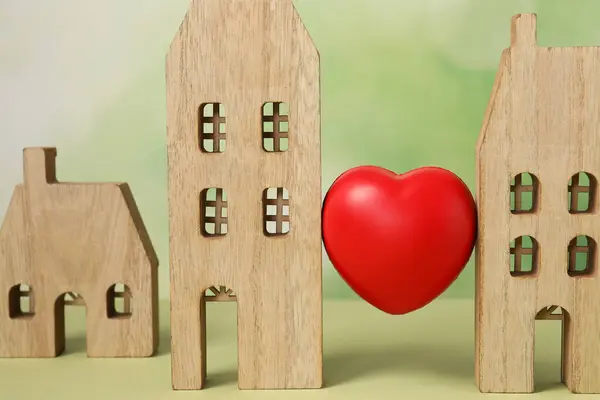 Long-distance relationship concept. Decorative heart between two wooden house models on light green background, closeup