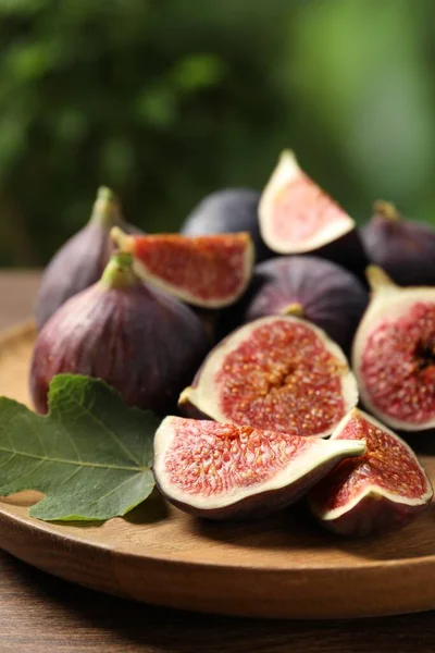 Whole and cut ripe figs with leaf on wooden table against blurred green background, closeup