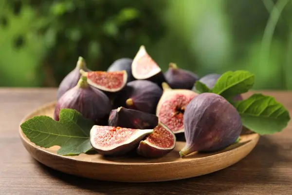 Whole and cut ripe figs with leaves on wooden table against blurred green background, closeup