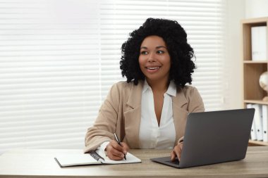 Mutlu kadın notlar yazıyor ve içerideki ahşap masada dizüstü bilgisayar kullanıyor. Metin için boşluk