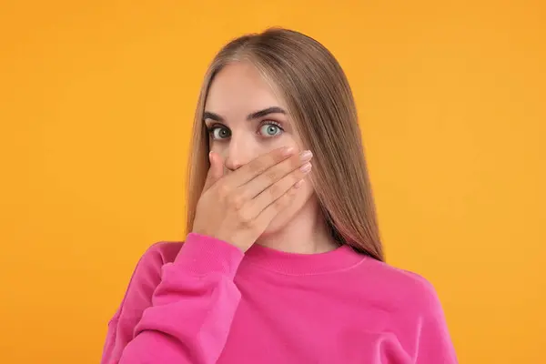 Embarrassed woman covering mouth with hand on orange background