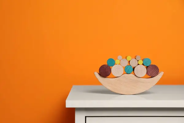 Wooden balance toy on white chest of drawers near orange wall, space for text. Children's development