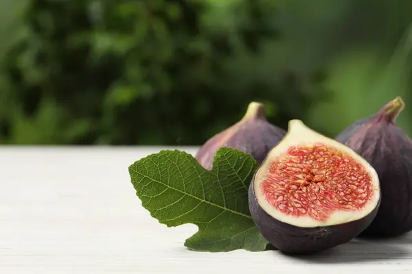 Whole and cut ripe figs with leaf on white wooden table against blurred green background, closeup. Space for text